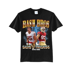 May include: Black t-shirt with a graphic of two football players in red and gold uniforms. The text "Bash Bros" is above the players, and "Sacks & Pancakes" is below them. The text "Kittle x Bosa" is below the players' names.