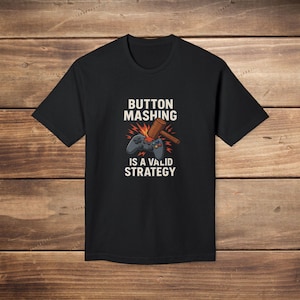 May include: Black t-shirt with the text "BUTTON MASHING IS A VALID STRATEGY" in white. The design includes a game controller and a wooden mallet. The shirt is made of a soft material.
