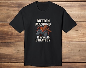 May include: Black t-shirt with the text "BUTTON MASHING IS A VALID STRATEGY" in white. The design includes a game controller and a wooden mallet. The shirt is made of a soft material.