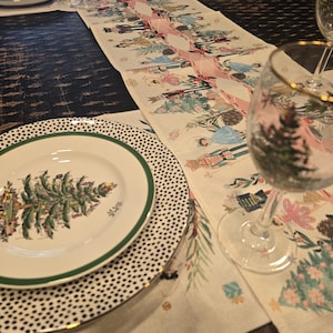 May include: A white table runner with a pink and green Christmas tree design. The runner is laid out on a dark wood table with two white plates with black polka dots and a green rim, a white plate with a green rim and a green Christmas tree design, and two wine glasses.