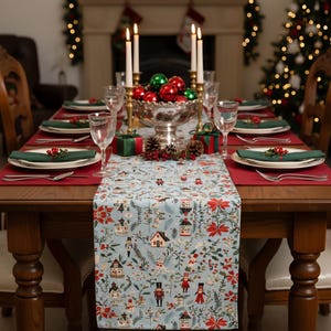 May include: A festive Christmas table setting with a light blue table runner featuring a winter village pattern. The table is set with red placemats, green napkins, and clear glassware. A silver bowl holds red and green ornaments, and candles add a warm glow.