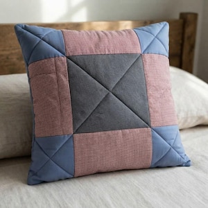 May include: A square throw pillow featuring a patchwork design with a central dark gray square, surrounded by four red and white gingham squares, and four blue corner squares. The entire surface is finished with a quilted diamond pattern.