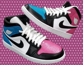 May include: Pair of high-top sneakers in black, white, blue, and pink. The sneakers feature a classic design with white laces, a black swoosh, and a white sole. The toe box and heel counter are pink, while the other shoe has a blue toe box.