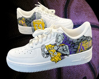 Puede incluir: Un par de zapatillas blancas con un diseño personalizado que presenta a un jugador de baloncesto vistiendo una camiseta morada y dorada con el número 24. Las zapatillas tienen un patrón de piel de serpiente negro y gris en el lateral y la palabra "Kobe" escrita en morado.