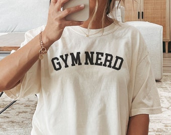 Puede incluir: Una camiseta blanca de gran tamaño con el texto "GYM NERD" impreso en negro. La modelo lleva la camiseta con pantalones cortos blancos y calcetines blancos con rayas negras.