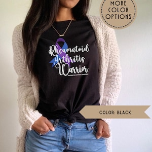 May include: A black t-shirt with a purple and blue ribbon design. The text "Rheumatoid Arthritis Warrior" is printed on the shirt. The hashtag "#rheumatoidarthritisawareness" is printed below the text.