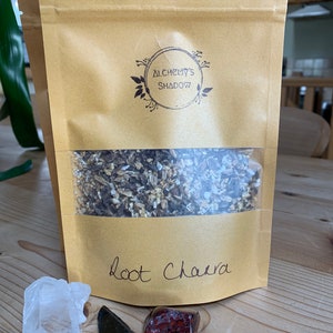 May include: A brown paper bag with a white label that says "Root Chaera" and a logo that says "Alchemy's Shadow". The bag contains dried herbs. There are three crystals next to the bag: a clear quartz crystal, a black obsidian arrowhead, and a red jasper stone.