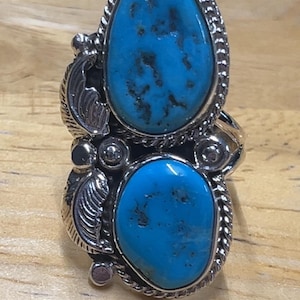 Pode incluir: Um anel de prata com duas pedras ovais de turquesa. As pedras azuis brilhantes apresentam padrões de matriz escura. O anel tem um design detalhado e texturizado com detalhes de folhas e corda.