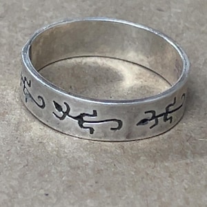 May include: A silver ring with etched, stylized symbols around the band. The ring has a simple, classic design and appears to be made of a light-colored metal. The symbols are dark and contrast with the silver color of the ring.