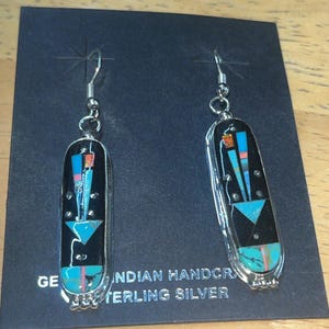 May include: A pair of handcrafted sterling silver earrings. Each earring features a black rectangular design with turquoise, blue, and orange geometric inlays. The text "INDIAN HANDCRAFTED STERLING SILVER" is visible on the backing.
