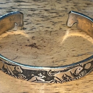 Puede incluir: Un brazalete de metal plateado con diseño abierto. La superficie exterior de la banda está decorada con un patrón en relieve de caballos y elementos florales sobre un fondo oscuro con textura hundida y detallada.