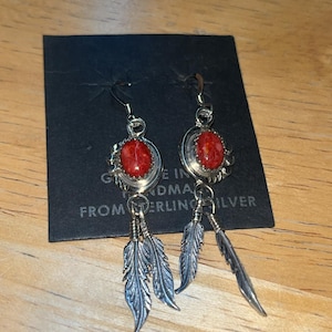 May include: Silver earrings with red oval stones and dangling feather charms. The earrings are on a black card with text that reads "Genuine Sterling Silver". The earrings are displayed on a wooden surface.