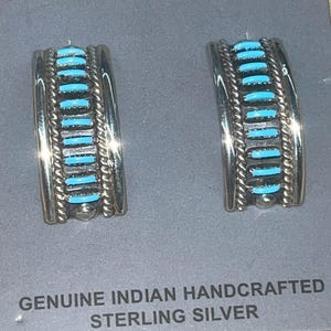 May include: A pair of silver earrings with turquoise inlay. The earrings are handcrafted and feature a geometric design. The text "GENUINE INDIAN HANDCRAFTED STERLING SILVER" is printed on the card.