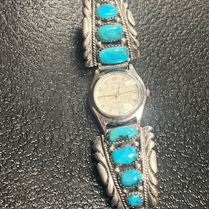 Op de afbeelding: Een zilveren horloge met een witte wijzerplaat en turquoise stenen op de band. De wijzerplaat van het horloge heeft zwarte Romeinse cijfers voor de uren en het woord "Quartz" staat erop gedrukt.