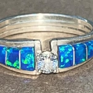 May include: Silver ring with a band of blue and green opal-like stones. A clear, round stone is set in the center. The ring has a unique design with a split band and a decorative stone setting.