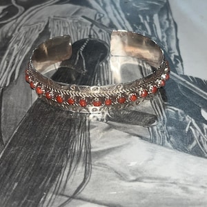 May include: A silver bracelet with a row of red coral beads. The bracelet has a decorative design with a repeating pattern.