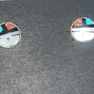 May include: A pair of silver earrings with turquoise, red, and black inlay. The earrings are round and have a silver backing.