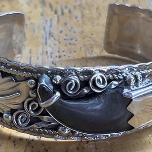 May include: A silver cuff bracelet featuring a central dark curved claw-like element, flanked by intricate metal leaf engravings, silver wire scrollwork, and small turquoise stones set at the ends of the open band design.