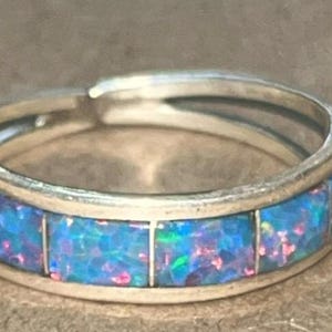 May include: A silver ring with a band of rectangular blue opal stones. The opal has flashes of pink and green. The ring is a simple band style, suitable for everyday wear.
