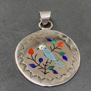 May include: A circular silver pendant featuring a bird and floral design. The bird is blue, with red, blue, and green details. The pendant has a decorative edge and a silver loop for a chain.