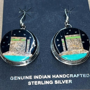May include: Pair of handcrafted sterling silver earrings. Each earring features a round, black background with a turquoise, tan, and green design. The earrings have a silver hook and are labeled "GENUINE INDIAN HANDCRAFTED STERLING SILVER."