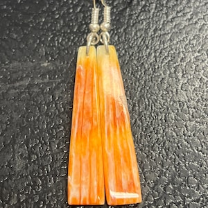 Peut inclure: Une paire de boucles d'oreilles pendantes avec des pendentifs en pierre rayés orange et blanc. Les boucles d'oreilles ont des crochets argentés.