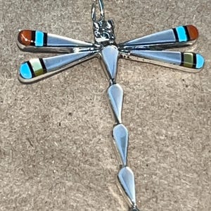 May include: A silver dragonfly pendant with turquoise, red, and yellow inlay. The dragonfly has a long, slender body and delicate wings.