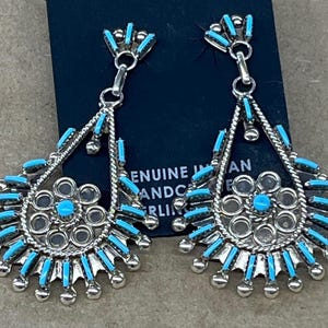 Pode incluir: Um par de brincos pendentes com um design turquesa e prata. Os brincos têm a forma de uma lágrima com um desenho de flor no centro. Os brincos são feitos de prata de lei e são adornados com pedras turquesa.