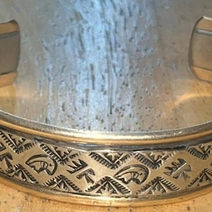 May include: A gold-coloured cuff bracelet featuring an engraved geometric pattern and bear motifs. The bracelet has a wide band with a detailed design around the centre. The open ends allow for easy wear. A classic piece of jewellery.