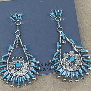 Pode incluir: Brincos pendurados prateados com detalhes turquesa. Cada brinco apresenta um formato de gota com um design floral central e barras turquesas. Os brincos são exibidos em um cartão azul.