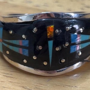 May include: A silver ring with a black band inlaid with turquoise, pink, and orange geometric designs. The ring has a polished silver finish and small silver accents. The ring is likely a piece of jewelry.