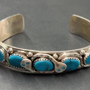 Op de afbeelding: Zilveren manchetarmband met vijf ovale turkooizen stenen. De armband heeft een gedetailleerd ontwerp met decoratieve elementen tussen de stenen. De turkooizen stenen zijn helderblauw, gezet in een zilveren band.