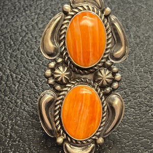 Könnte beinhalten: Ein Ring aus Sterlingsilber mit zwei ovalen, orangefarbenen Spiny-Oyster-Muschelsteinen. Der Ring hat ein detailliertes Design mit einem seilartigen Rand um die Steine.