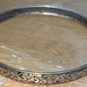 May include: A silver-toned bangle bracelet with an intricate floral pattern around the band. The bracelet is circular and appears to be made of metal. The design is repeated around the entire circumference of the bracelet.
