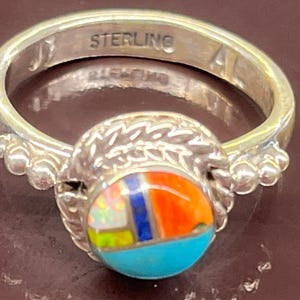 May include: Sterling silver ring with a colorful inlaid stone. The stone features turquoise, orange, blue, and white sections. The ring band is engraved with "STERLING". The ring has a decorative design around the stone.