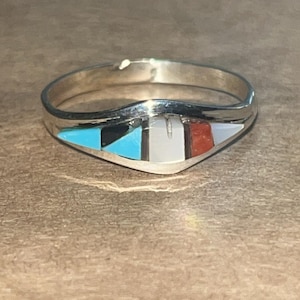 May include: A silver ring with a triangular inlay featuring turquoise, black, white, and red geometric shapes. The ring has a smooth, polished band and a decorative design on the front.