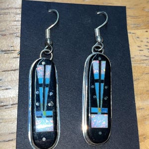 May include: A pair of silver-toned earrings with a black background. Each earring features a rectangular design with a black base, accented with blue, white, and iridescent details. The earrings have a hook closure.