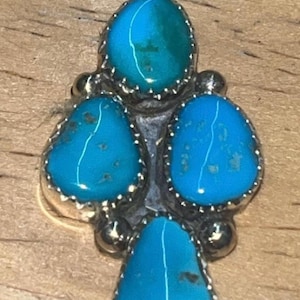 May include: A silver pendant featuring four turquoise stones. The stones are teardrop and oval shaped, set in a silver bezel with a beaded edge. The pendant has a bail at the top for a chain.