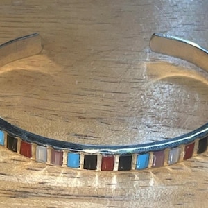 May include: Silver-tone cuff bracelet with a row of colorful rectangular inlays. The inlays include turquoise, black, red, white, and pink. The bracelet is open-ended and rests on a wooden surface.