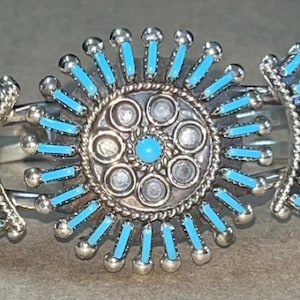 Pode incluir: Pulseira de punho de prata com um design central de explosão solar e detalhes turquesa. A pulseira apresenta barras cravejadas de turquesa que irradiam de um medalhão central, criando um efeito visual marcante. É uma joia.