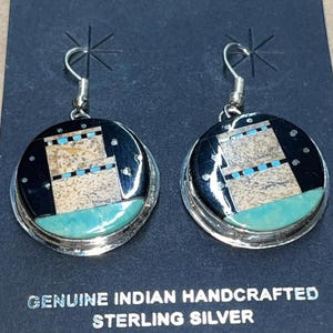 May include: Pair of handcrafted sterling silver earrings. Each earring features a circular design with a black background, a turquoise lower section, and a central geometric pattern in beige and blue. The earrings have a hook closure.