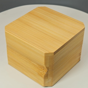 May include: A square, light-colored bamboo box with a beveled top and sides. The box is made of multiple bamboo strips, and the grain of the wood is visible. The box is sitting on a white surface.