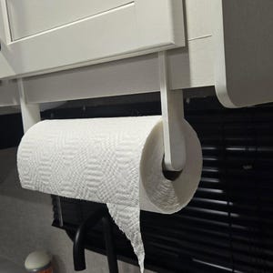 May include: White paper towel holder mounted under a white cabinet. The holder is made of white plastic and has a simple design. A roll of white paper towels is attached to the holder.