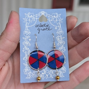 May include: A pair of round earrings with a red and blue segmented design. The earrings have a silver-colored metal frame and a small gold bead at the bottom. The earrings are displayed on a light blue card with the text "eclectic grace".