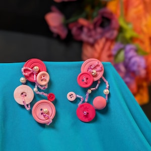 May include: A pair of handmade button earrings in shades of pink, featuring various sizes and textures. The earrings are adorned with small beads and pearls, set against a teal background.
