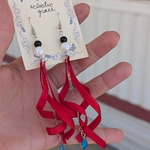 Puede incluir: Pendientes colgantes en una tarjeta de presentación color crema con la etiqueta 'eclectic grace'. Cada pendiente tiene un gancho plateado, una cuenta negra, una cuenta blanca, una cinta roja en espiral, una cadena plateada y un dije de gota azul.