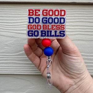 May include: A retractable badge reel with the text "BE GOOD DO GOOD GOD BLESS GO BILLS" in red and blue. The reel has a red and blue bead design. The badge reel is held in a hand.