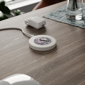 May include: A white wireless charger with a black and white image of a crow perched on a branch in front of a full moon. A white charging cable and a white charging case are also visible on a wooden surface.