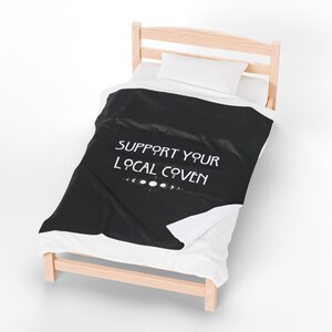 May include: A black blanket with the text "SUPPORT YOUR LOCAL COVEN" in white, along with a series of moon phase symbols. The blanket is displayed on a bed with a white pillow and sheets, and a light wood frame.