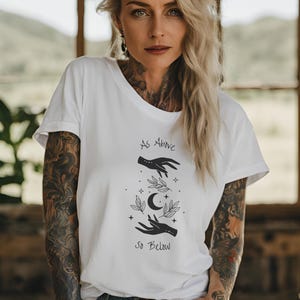 May include: White t-shirt with a black graphic design of two hands reaching towards a crescent moon with stars and leaves. The text "As Above So Below" is printed below the graphic.